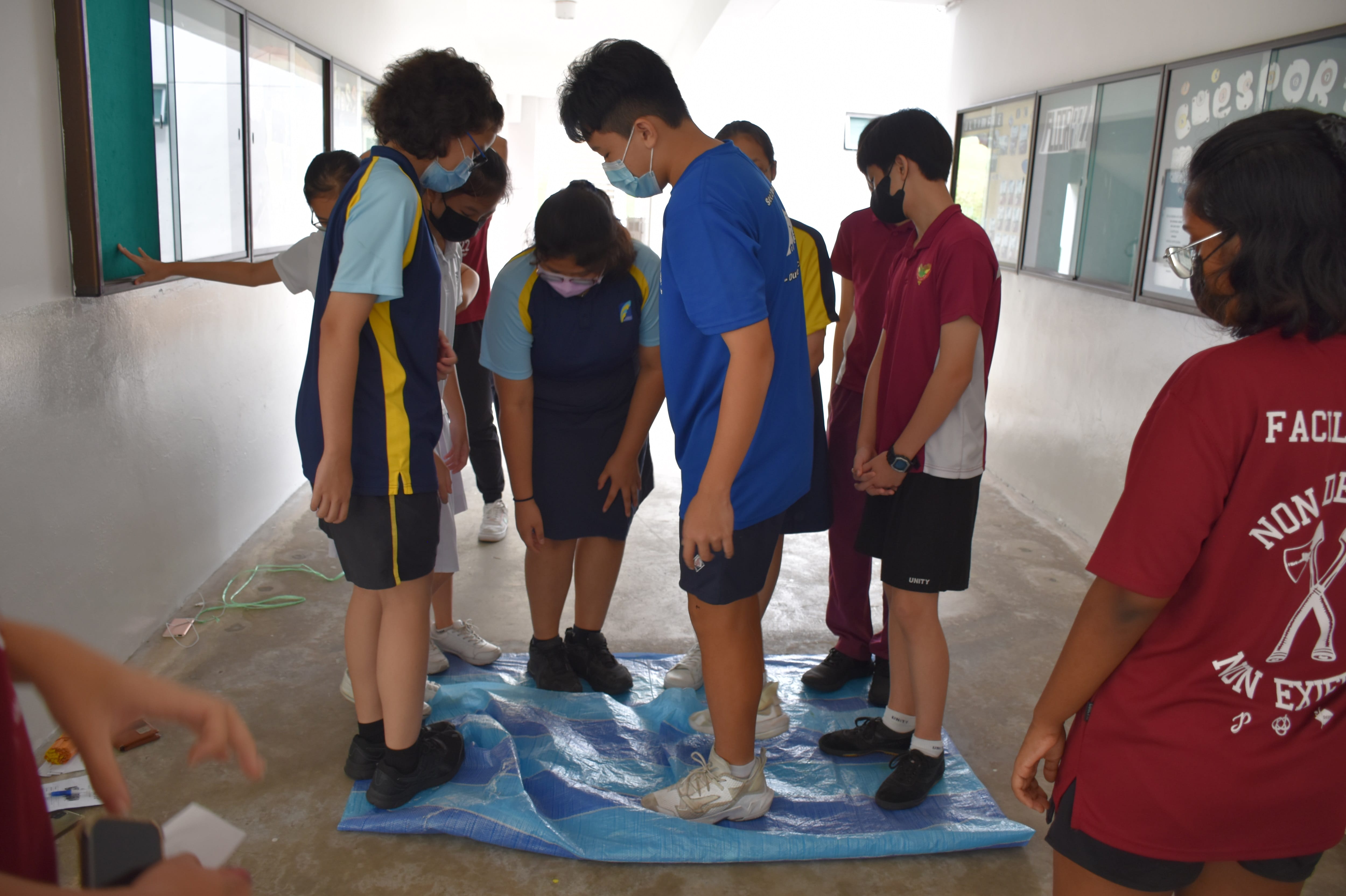Students wearing face masks stand on a blue tarp on a school corridor floor, looking down at it.